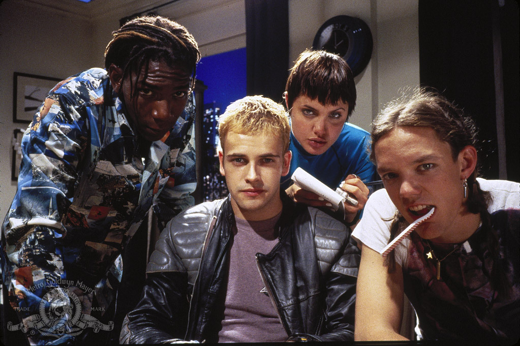 Uma foto promocional do filme Hackers: Piratas de Computador, mostrando quatro de seus cinco protagonistas encarando diretamente a câmera. Todos vestem roupas casual-alternativas típicas da segunda metade dos anos 90, a época das grandes raves e dos primórdios da internet. No canto inferior direito da imagem, está a marca d'água da produtora do filme, a MGM Studios (atualmente Amazon Studios).