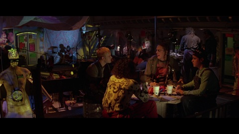 Uma captura de tela do filme Hackers: Piratas de Computador que mostra Dade Murphy, Lord Nikon, Emmanuel "Cereal Killer" Goldstein (Matthew Lillard) e Joey comendo um lanche de fast-food juntos na tumultuada e collorida rave Cyberdelia.